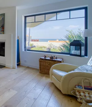 Un fauteuil beige repose près d'une grande fenêtre donnant sur la mer, entouré d'une cheminée, d'une petite table en osier et de livres.