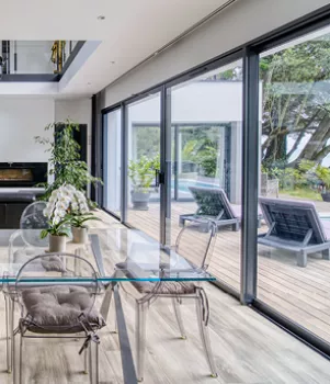 Salle à manger moderne avec table en verre, donnant sur une terrasse en bois avec des chaises longues et vue sur le jardin arboré, grâce à de grandes baies vitrées.