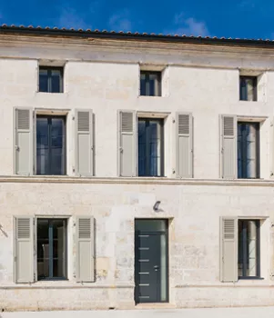 Maison en pierre à volets ouverts, exposée au soleil, située dans un environnement résidentiel calme avec ciel bleu en arrière-plan.