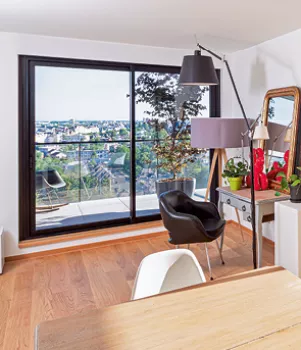 Salon moderne accueillant un fauteuil noir près d’une baie vitrée avec vue panoramique, meublé d’une lampe sur pied et d’une table avec des plantes vertes.