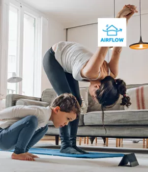 Femme fait des exercices de yoga tandis qu'un enfant l'observe attentivement, tout ceci dans un salon bien éclairé avec un canapé et une lampe suspendue.