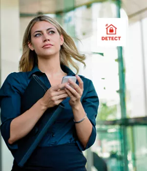 Une femme utilise son smartphone en marchant devant un bâtiment moderne avec des façades vitrées. Elle tient également un dossier sous son bras.
