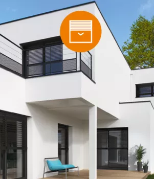 Maison moderne avec façade blanche. Volets roulants semi-fermés. Terrasse avec un banc. Fenêtres noires contrastant avec les murs. Arbre visible en arrière-plan sous un ciel bleu.