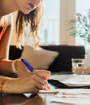 Une femme écrit avec un stylo bleu sur des documents étalés sur une table, dans une pièce lumineuse avec des fenêtres en arrière-plan.
