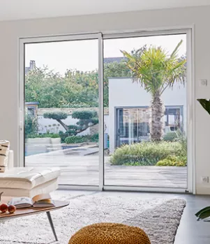 Une grande baie vitrée donne sur un jardin ensoleillé avec des palmiers, visible depuis un salon moderne et lumineux.