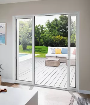 Une baie vitrée coulissante donne sur une terrasse en bois, meublée d'un canapé avec des coussins colorés, et encadrée par un jardin verdoyant.