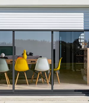 Quatre chaises colorées entourent une table en bois, visibles à travers une grande baie vitrée, surplombées par un volet roulant gris.