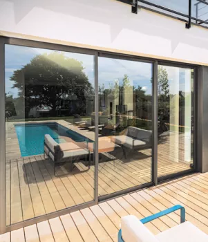 Porte-fenêtre vitrée, reflétant la terrasse en bois avec piscine entourée de meubles de jardin, dans un environnement extérieur verdoyant.