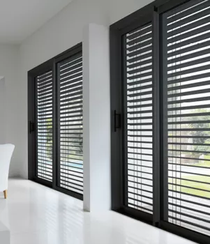 Brise soleil orientable. Des stores vénitiens en aluminium noir couvrent de grandes baies vitrées, laissant filtrer la lumière du jour dans une pièce claire et moderne avec un fauteuil blanc.