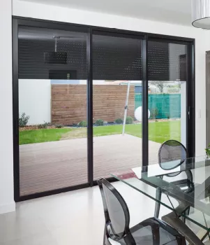 Baie vitrée dans une salle à manger avec table en verre et chaises transparentes, donnant sur une terrasse et un jardin clôturé.