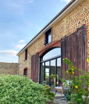 Grande bâtisse en pierre rénovée avec baies vitrées et portes en bois, entourée de verdure sous un ciel bleu.