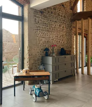 Une petite voiture en métal est stationnée devant une table basse en bois, dans une pièce avec des murs en pierre et une commode grise.