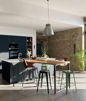 Cuisine moderne avec îlot central, des tabourets, plantes, et luminaires suspendus. Contexte lumineux et spacieux avec mur en pierre apparente.