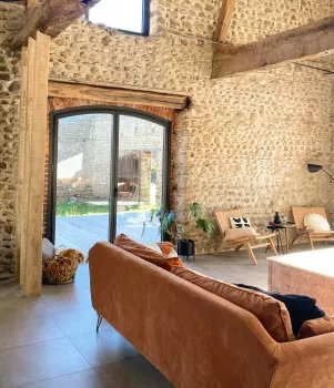 Canapé en cuir marron, situé dans un salon lumineux avec murs en pierres apparentes, grandes fenêtres et mobilier moderne. Plan de travail, plantes et chaise en bois complètent l'espace convivial.