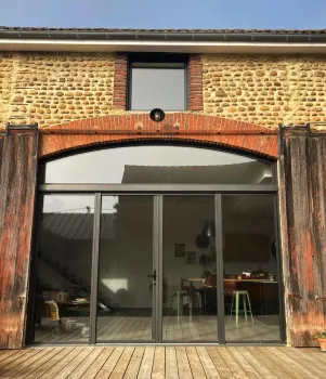 Grande porte vitrée réfléchissante donnant sur une terrasse en bois, encadrée de murs en pierre ancienne et de volets en bois. L'intérieur révèle une cuisine moderne.