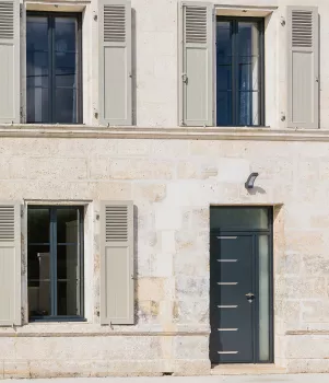 Façade de maison en pierre aux volets gris fermés, avec porte noire centrale, façade claire, lampadaire, garage à gauche.