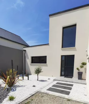 Maison moderne en façade crème, large fenêtre, porte noire, petit jardin de graviers blancs, plantes décoratives, allée avec dalles grises, ciel bleu.