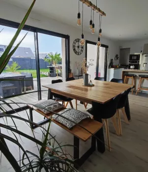 Table en bois garnie de coussins, entourée de chaises noires, dans une salle à manger lumineuse ouverte sur un jardin grâce à des baies vitrées.