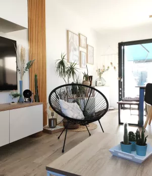 Un fauteuil moderne avec coussin repose dans un salon lumineux, entouré de plantes vertes, devant une télévision et une grande porte vitrée.