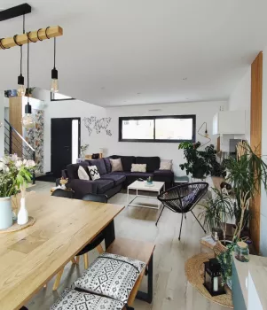 Salon moderne et épuré, un grand canapé gris autour d'une table basse, plantes vertes disposées, table en bois avec décoration florale, éclairage suspendu, ambiance lumineuse et accueillante.