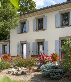 Maison blanche à volets gris entourée de fleurs colorées et de buissons, située dans un jardin ensoleillé avec un banc en bois devant.