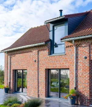 Maison en briques rouges, avec grandes fenêtres et un toit en tuiles, située dans un jardin verdoyant sous un ciel légèrement nuageux.
