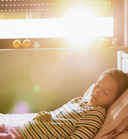 Une fille dort paisiblement, allongée sur un lit, baignée par la lumière du soleil matinal qui entre par la fenêtre de sa chambre.