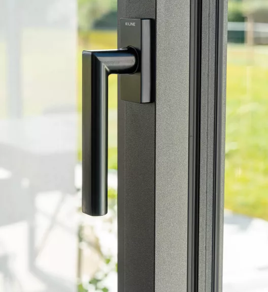 Poignée noire est fixée sur une porte-fenêtre grise, avec une vue lumineuse sur un jardin en arrière-plan.