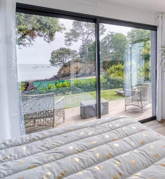 Chambre avec grande baie vitrée ouverte sur une terrasse en bois, offrant vue sur un jardin verdoyant, des arbres et la mer avec des bateaux au loin.