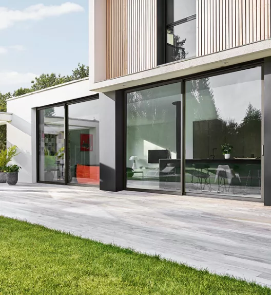 Maison moderne avec grandes baies vitrées donnant sur une terrasse en bois, entourée de gazon vert, sous un ciel bleu clair.