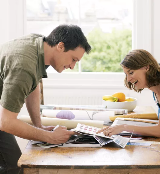 Deux personnes consultent des échantillons de couleurs sur une table en bois, souriant et discutant, dans une pièce lumineuse avec une fenêtre et un bol de fruits.