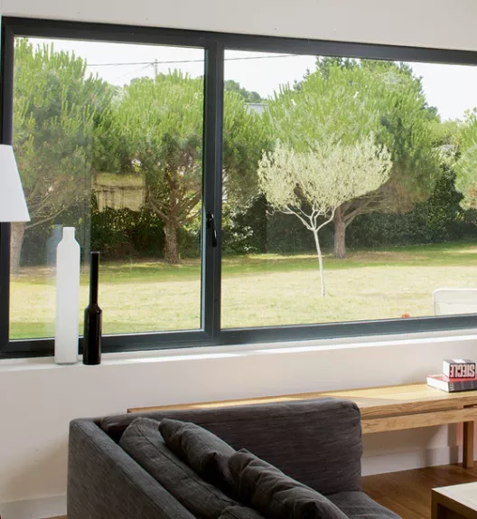 Un canapé gris est placé dans un salon lumineux avec une grande fenêtre qui donne sur un jardin verdoyant.