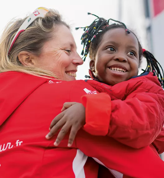 Une femme en veste rouge enlace et sourit à une fillette joyeuse aux cheveux tressés, dans un environnement extérieur.