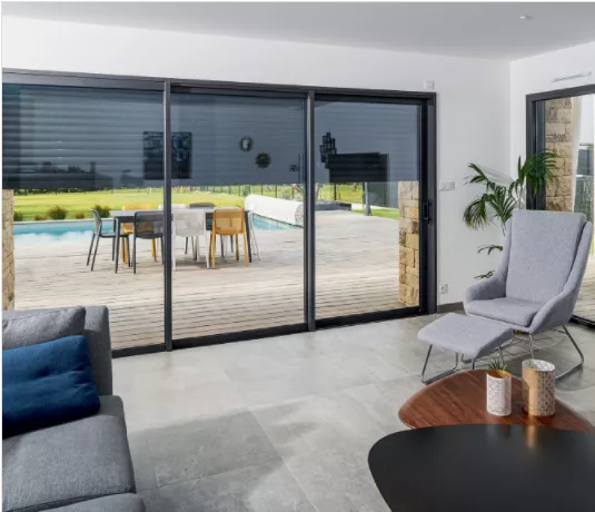 Un salon moderne avec canapé et fauteuils donne sur une terrasse en bois équipée de table et chaises, près d'une piscine extérieure.