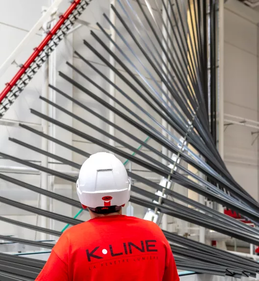 Un ouvrier en casque et t-shirt rouge observe une machine avec des pièces métalliques en forme d'éventail dans une usine.