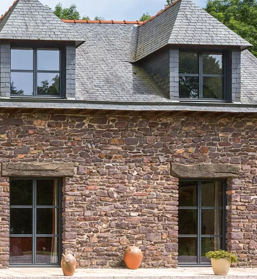 Maison en pierres, avec toit en ardoises et deux fenêtres en pignon, ornée de trois pots en terre cuite sur la terrasse devant.