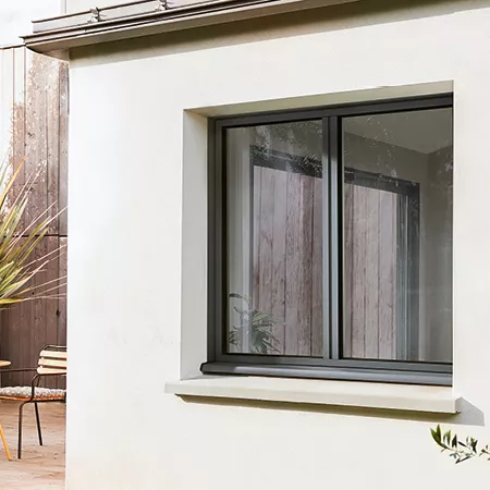 Fenêtre coulissante, fermée, encastrée dans un mur blanc, avec un patio en arrière-plan contenant une chaise et une plante.