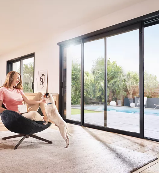 Personne assise caressant un chien, dans un salon lumineux avec de grandes baies vitrées donnant sur une terrasse avec piscine et jardin verdoyant.