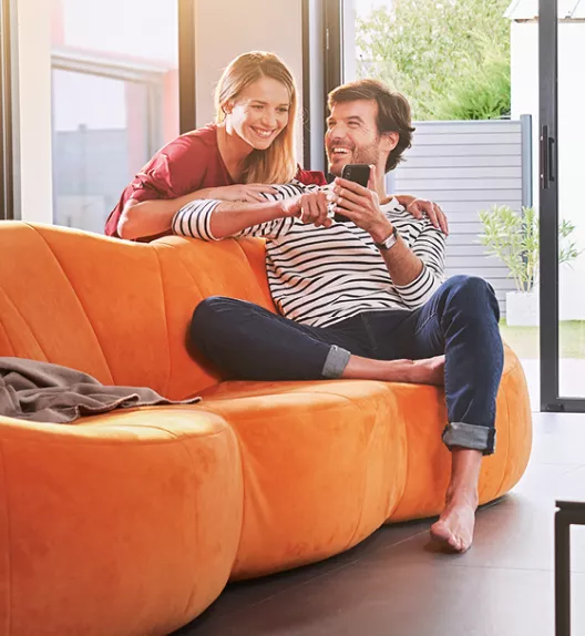 Un couple assis sur un canapé orange relaxe en regardant un smartphone, dans un salon lumineux avec une grande baie vitrée.