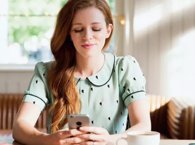La femme utilise son téléphone portable tout en étant assise dans un café lumineux avec une tasse de café à côté d'elle.