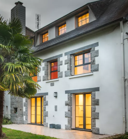 La maison à trois étages et fenêtres illuminées se trouve dans un jardin avec un palmier à gauche, créant une ambiance chaleureuse et accueillante en soirée.