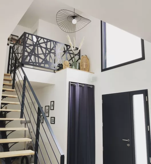 Une cage d'escalier design en bois et métal monte vers une mezzanine avec des plantes et une décoration moderne, éclairée par une grande fenêtre et un lustre contemporain.