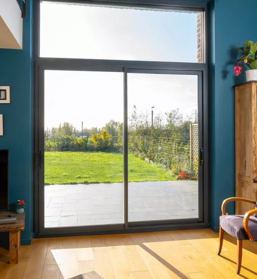Grande baie vitrée coulissante illuminant un salon avec parquet en bois, ouvrant sur un jardin verdoyant et ensoleillé entouré de buissons et de fleurs.