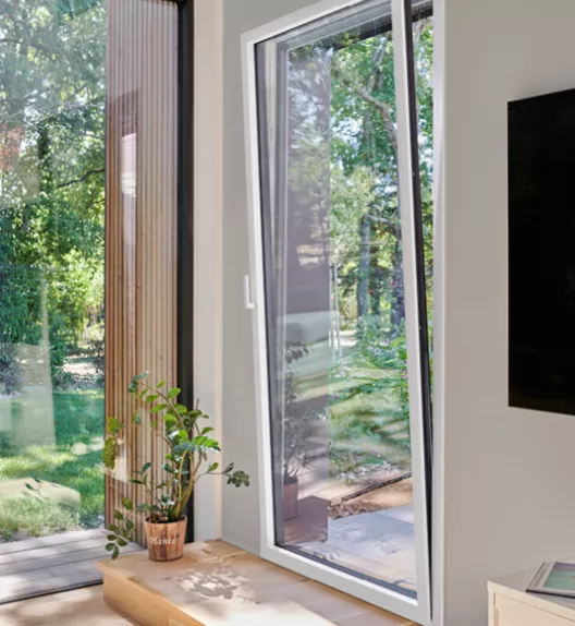 Fenêtre basculée laissant entrer la lumière, avec un pot de plante à côté, donnant sur un jardin verdoyant.