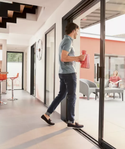 Homme portant une chemise bleue, tenant un arrosoir rouge, marche vers la terrasse où une femme est assise sur une chaise en extérieur d'une maison moderne.