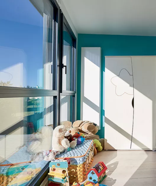 Des jouets en peluche reposent près d'une fenêtre lumineuse, dans une chambre d'enfant avec une armoire blanche et un mur turquoise.