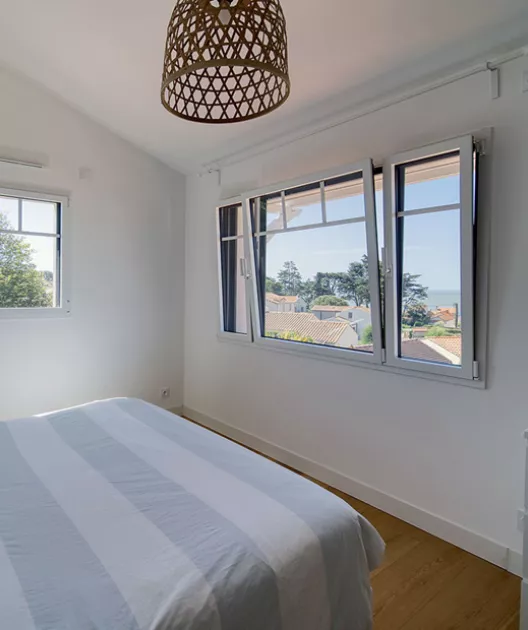 Un lit avec une housse rayée repose dans une chambre lumineuse, surplombée par un luminaire suspendu, avec des fenêtres ouvertes offrant une vue dégagée sur un paysage extérieur.