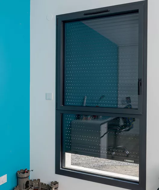 Fenêtre noire avec volet roulant, située dans une pièce à mur bleu, donnant sur un bureau avec chaise et objets divers.