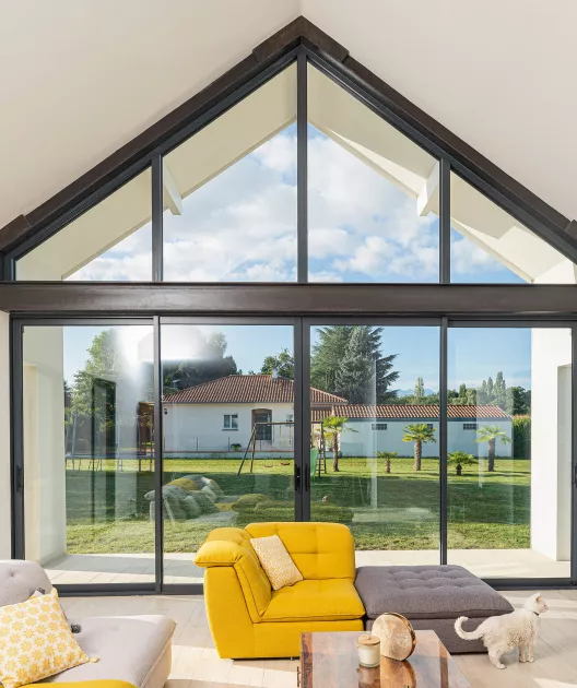 Canapé jaune lumineux et chat jouent dans un salon spacieux avec une grande baie vitrée donnant sur un jardin ensoleillé et une maison de style méditerranéen.