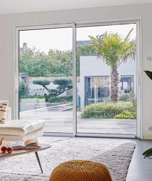 Une maison moderne avec une grande baie vitrée s'ouvre sur un jardin verdoyant avec une terrasse en bois, une palmeraie et des buissons.